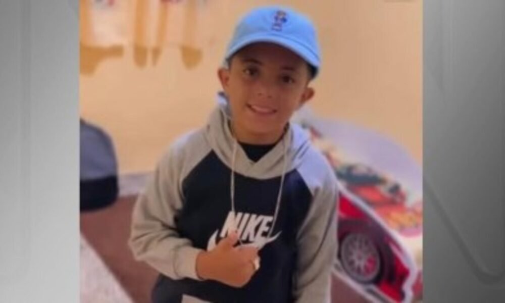 Smiling young boy wearing a blue cap and a gray-black Nike hoodie, looking at the camera indoors.