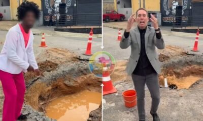 Two people stand around a large pothole with muddy water on a city street orange traffic cones marking the area A person in a gray suit gestures while speaking a woman in pink pants watches on the left