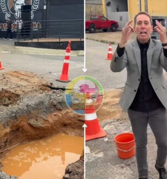 Two people stand around a large pothole with muddy water on a city street orange traffic cones marking the area A person in a gray suit gestures while speaking a woman in pink pants watches on the left