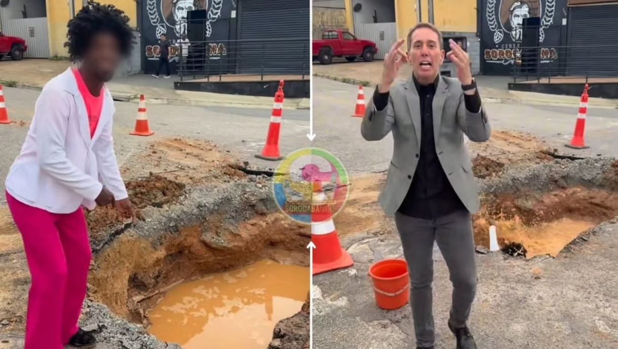 Two people stand around a large pothole with muddy water on a city street orange traffic cones marking the area A person in a gray suit gestures while speaking a woman in pink pants watches on the left