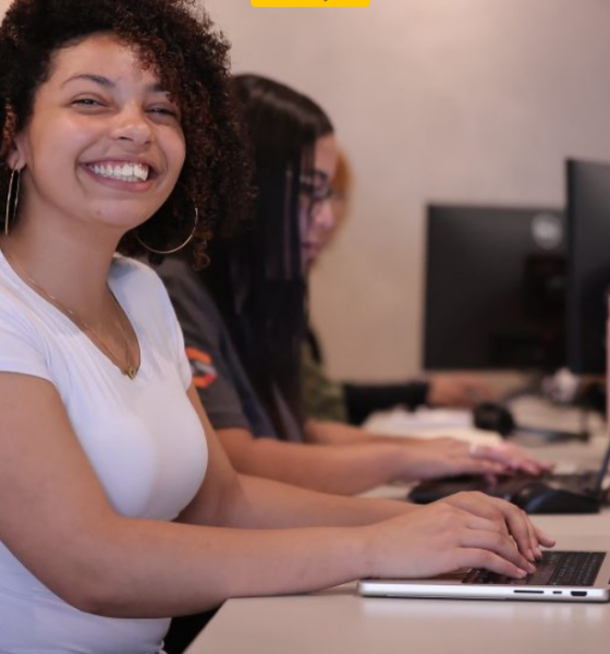 Smiling woman with curly hair in a white shirt sits at a computer desk typing on a laptop in a row of coworkers at PCs