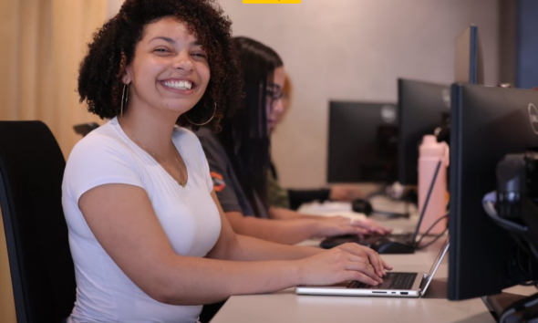 Smiling woman with curly hair in a white shirt sits at a computer desk typing on a laptop in a row of coworkers at PCs