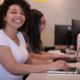 Smiling woman with curly hair in a white shirt sits at a computer desk typing on a laptop in a row of coworkers at PCs