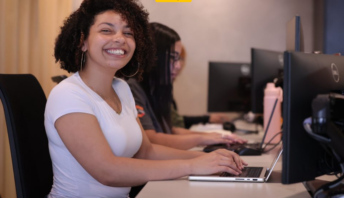 Smiling woman with curly hair in a white shirt sits at a computer desk typing on a laptop in a row of coworkers at PCs