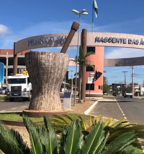Entrance arch to Pilar do Sul with a large stone pedestal sculpture in a circular planter and palm plants in the foreground cars on the street behind
