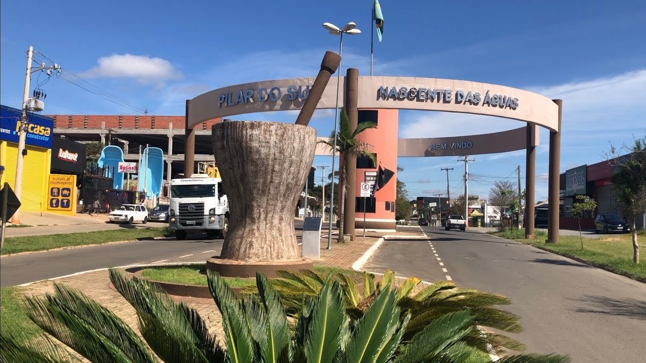 Entrance arch to Pilar do Sul with a large stone pedestal sculpture in a circular planter and palm plants in the foreground cars on the street behind