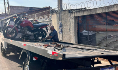 Tow truck carrying a red motorcycle with a man securing it on the flatbed in a street scene behind a barbed wire fence