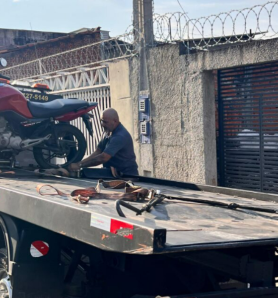 Tow truck carrying a red motorcycle with a man securing it on the flatbed in a street scene behind a barbed wire fence
