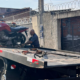 Tow truck carrying a red motorcycle with a man securing it on the flatbed in a street scene behind a barbed wire fence