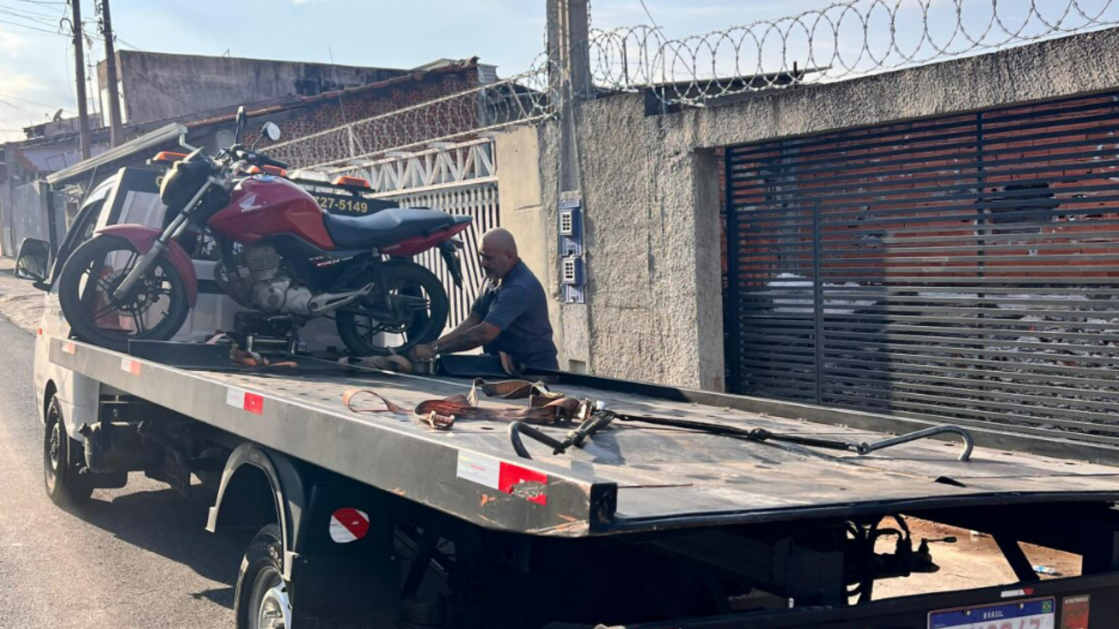 Tow truck carrying a red motorcycle with a man securing it on the flatbed in a street scene behind a barbed wire fence