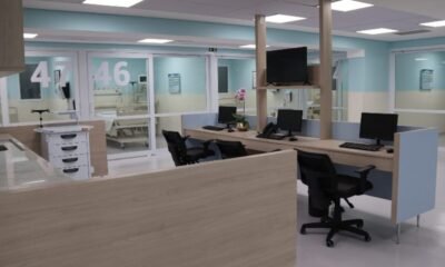 Hospital reception desk with multiple workstations and computer monitors in a bright modern ward area glass doors labeled  and  in the background