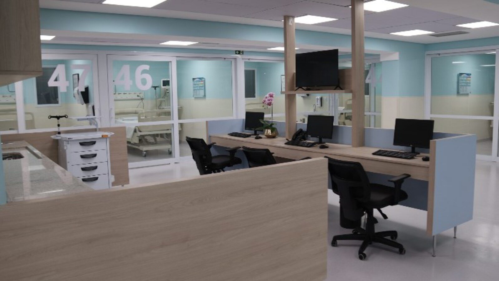 Hospital reception desk with multiple workstations and computer monitors in a bright modern ward area glass doors labeled  and  in the background