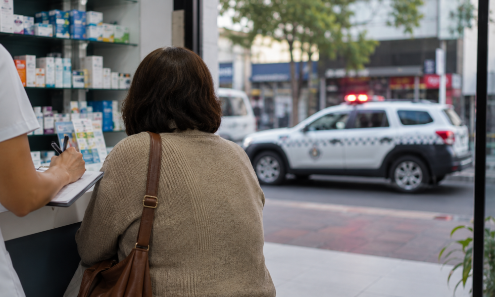 Customer with a brown shoulder bag at a store counter writes on a clipboard while a pharmacist assists, shelves of medications behind them, and a police SUV with flashing lights visible outside the window.