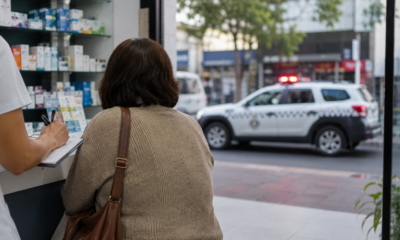 Customer with a brown shoulder bag at a store counter writes on a clipboard while a pharmacist assists shelves of medications behind them and a police SUV with flashing lights visible outside the window