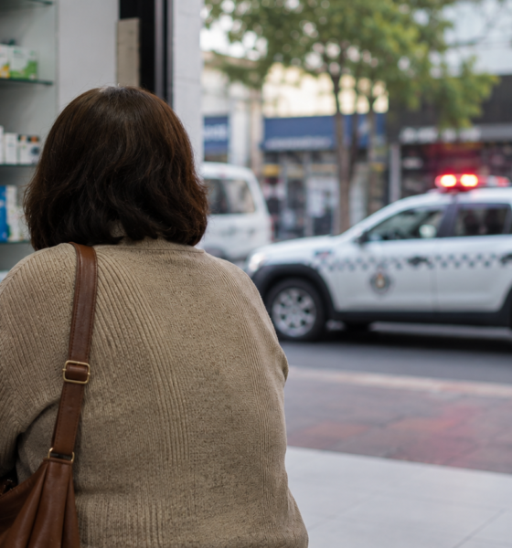 Customer with a brown shoulder bag at a store counter writes on a clipboard while a pharmacist assists shelves of medications behind them and a police SUV with flashing lights visible outside the window