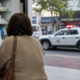 Customer with a brown shoulder bag at a store counter writes on a clipboard while a pharmacist assists shelves of medications behind them and a police SUV with flashing lights visible outside the window