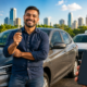 Smiling man in a blue shirt holds up car key beside a gray sedan with a taxi waiting in the background