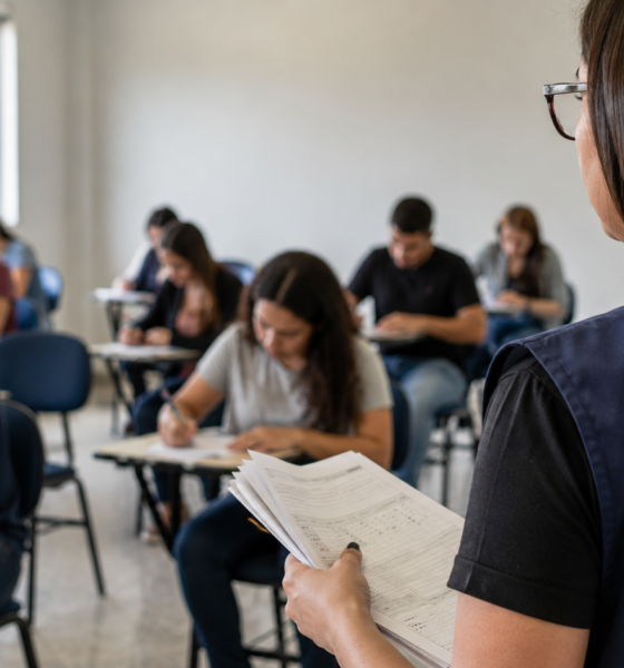 Instructor at the front of a classroom holds papers while students sit at desks and take notes nearby.