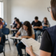 Instructor at the front of a classroom holds papers while students sit at desks and take notes nearby