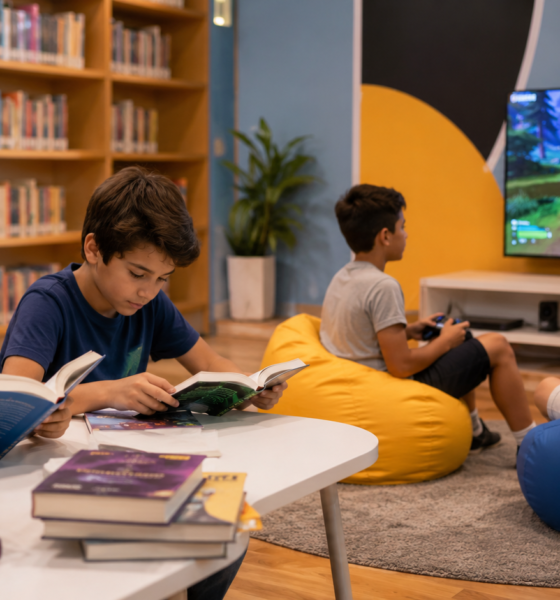 Children reading books at a table in a library while two others play a video game on a large screen in the background