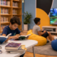 Children reading books at a table in a library while two others play a video game on a large screen in the background