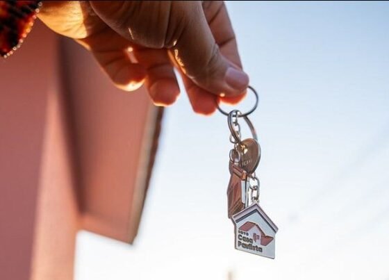 Person holding a keyring with a small house shaped keychain against a clear sky suggesting a new home or property handover