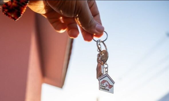 Person holding a keyring with a small house shaped keychain against a clear sky suggesting a new home or property handover