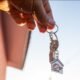 Person holding a keyring with a small house shaped keychain against a clear sky suggesting a new home or property handover