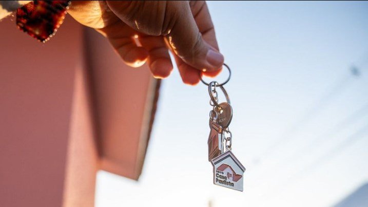 Person holding a keyring with a small house shaped keychain against a clear sky suggesting a new home or property handover
