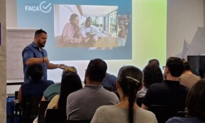 Male presenter in a blue shirt gestures toward a projected slide showing people collaborating at a table with a blue and lime green background and an audience seated facing forward
