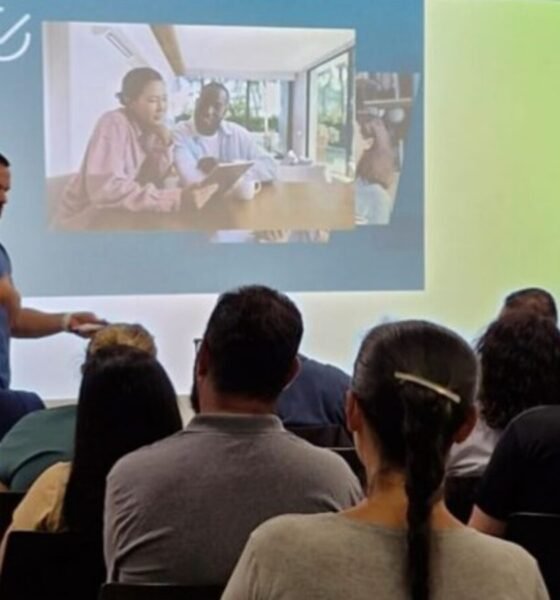 Male presenter in a blue shirt gestures toward a projected slide showing people collaborating at a table with a blue and lime green background and an audience seated facing forward