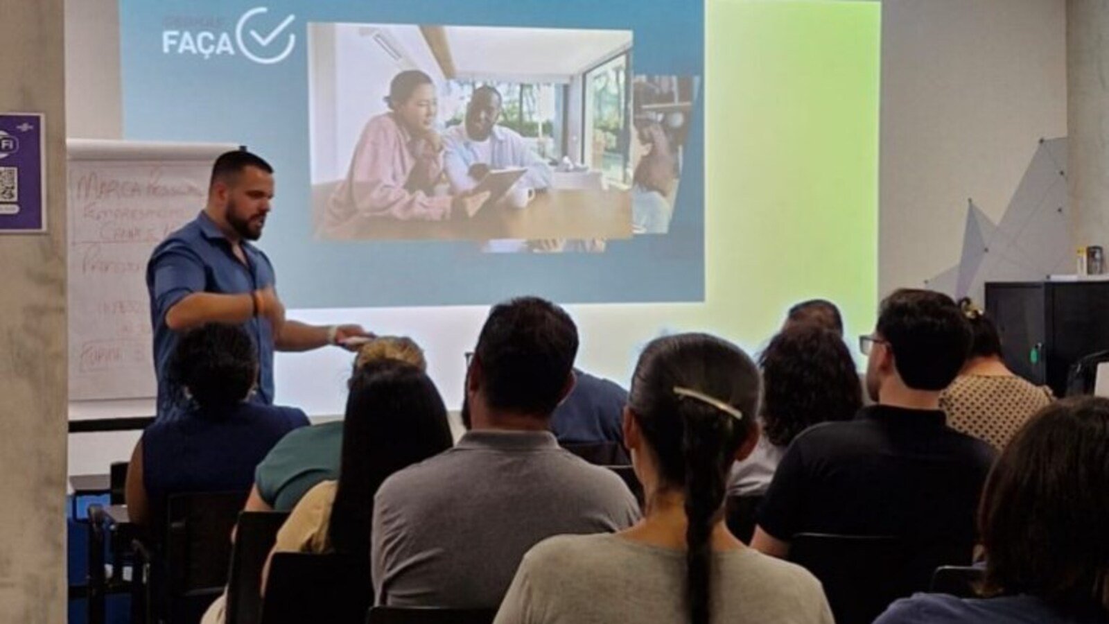 Male presenter in a blue shirt gestures toward a projected slide showing people collaborating at a table with a blue and lime green background and an audience seated facing forward