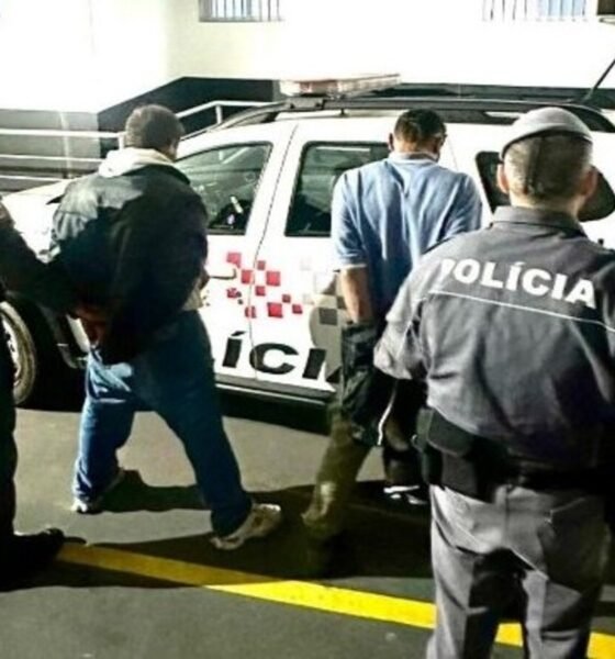 A man is being escorted by multiple police officers toward a white police SUV in a dimly lit parking area by a railing