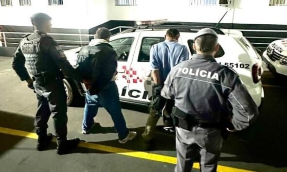 A man is being escorted by multiple police officers toward a white police SUV in a dimly lit parking area by a railing