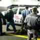 A man is being escorted by multiple police officers toward a white police SUV in a dimly lit parking area by a railing