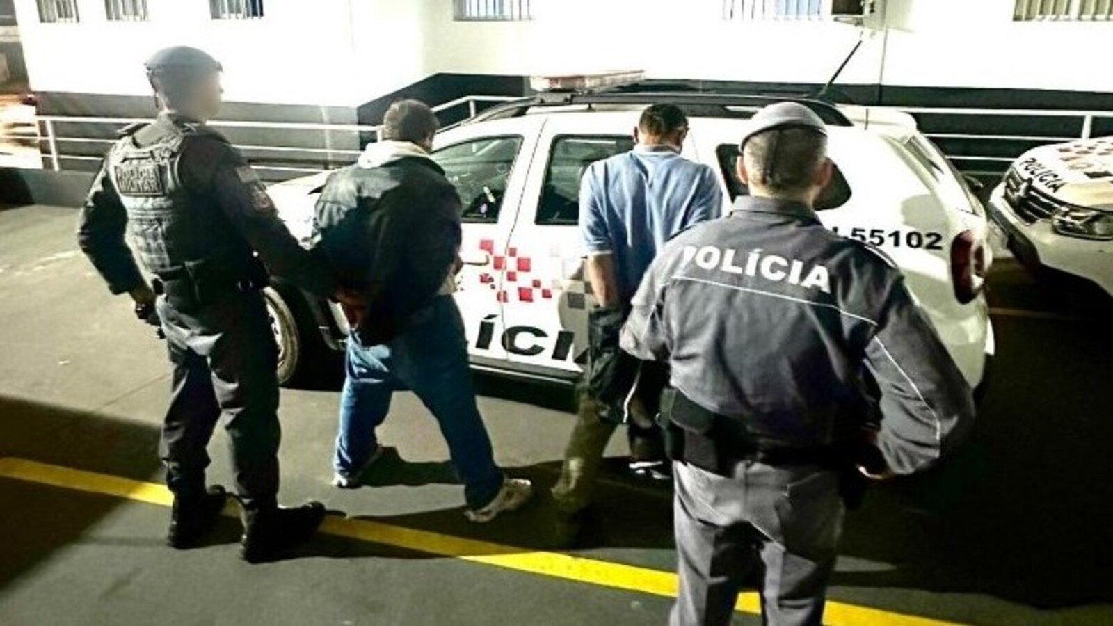 A man is being escorted by multiple police officers toward a white police SUV in a dimly lit parking area by a railing