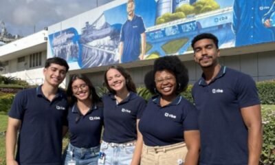 Five smiling young adults in navy polo shirts with a company logo stand together outdoors in front of a mural.