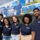 Five smiling young adults in navy polo shirts with a company logo stand together outdoors in front of a mural