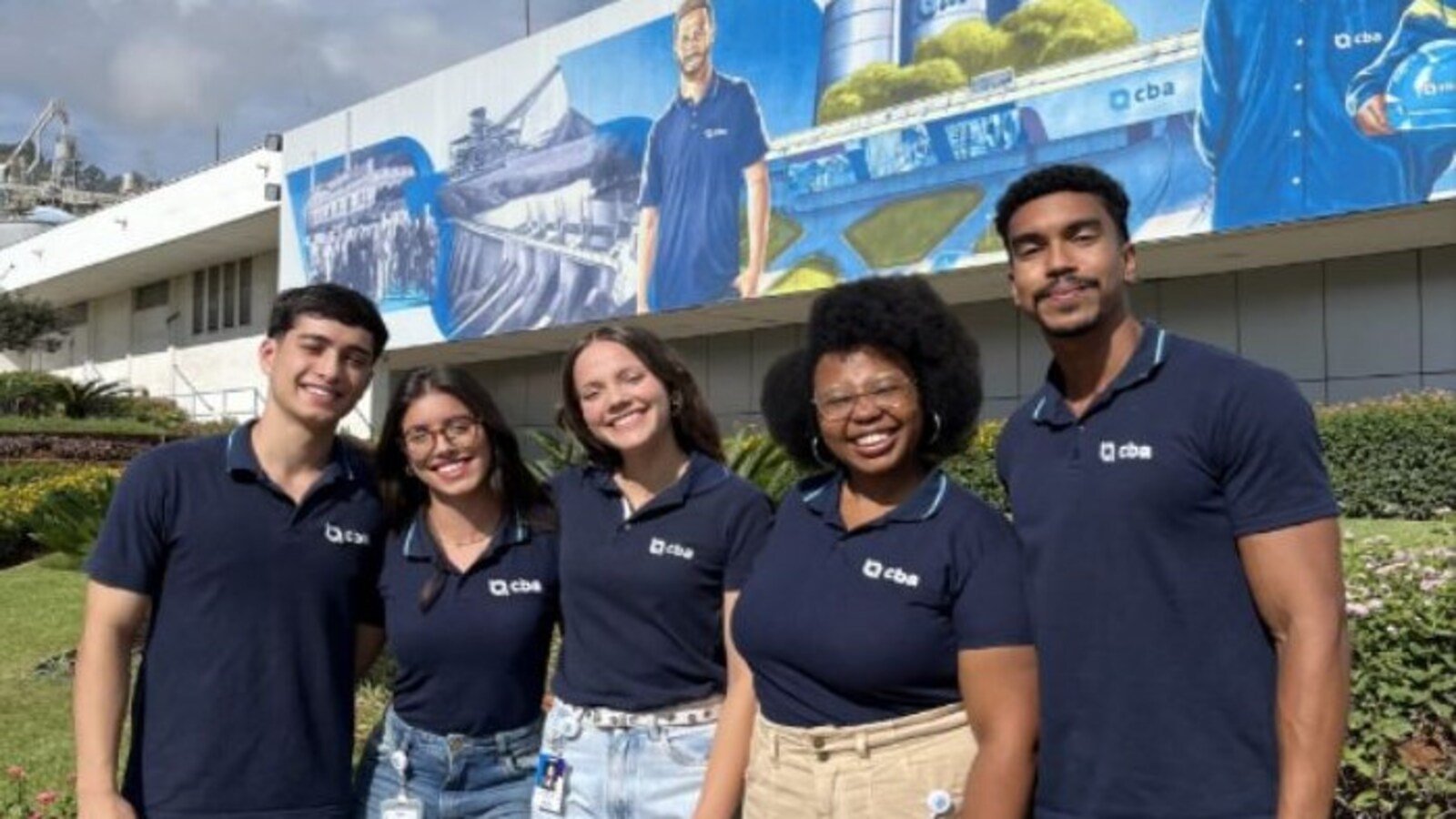 Five smiling young adults in navy polo shirts with a company logo stand together outdoors in front of a mural
