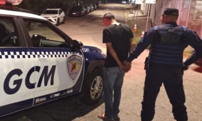 Uniformed officer detains a man in a black tshirt and jeans handcuffing him beside a white and blue GCM police SUV on a dim city street at night