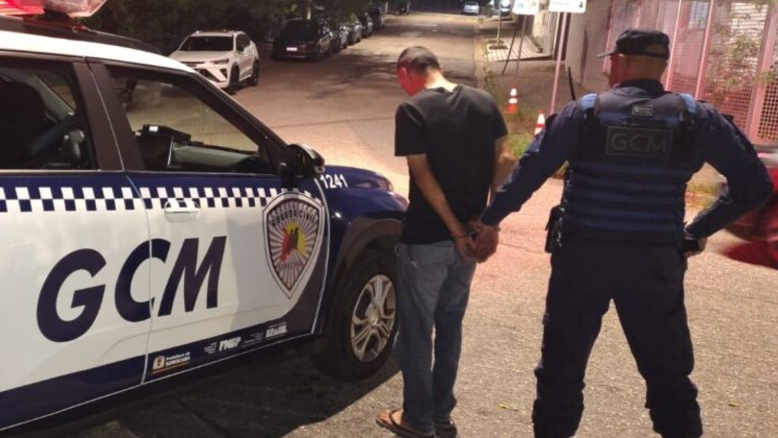 Uniformed officer detains a man in a black tshirt and jeans handcuffing him beside a white and blue GCM police SUV on a dim city street at night