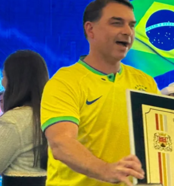 Man in a yellow Brazil jersey holds a large framed certificate on stage with a Brazilian flag in the background