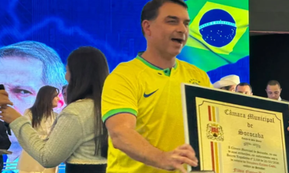 Man in a yellow Brazil jersey holds a large framed certificate on stage with a Brazilian flag in the background