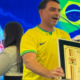 Man in a yellow Brazil jersey holds a large framed certificate on stage with a Brazilian flag in the background