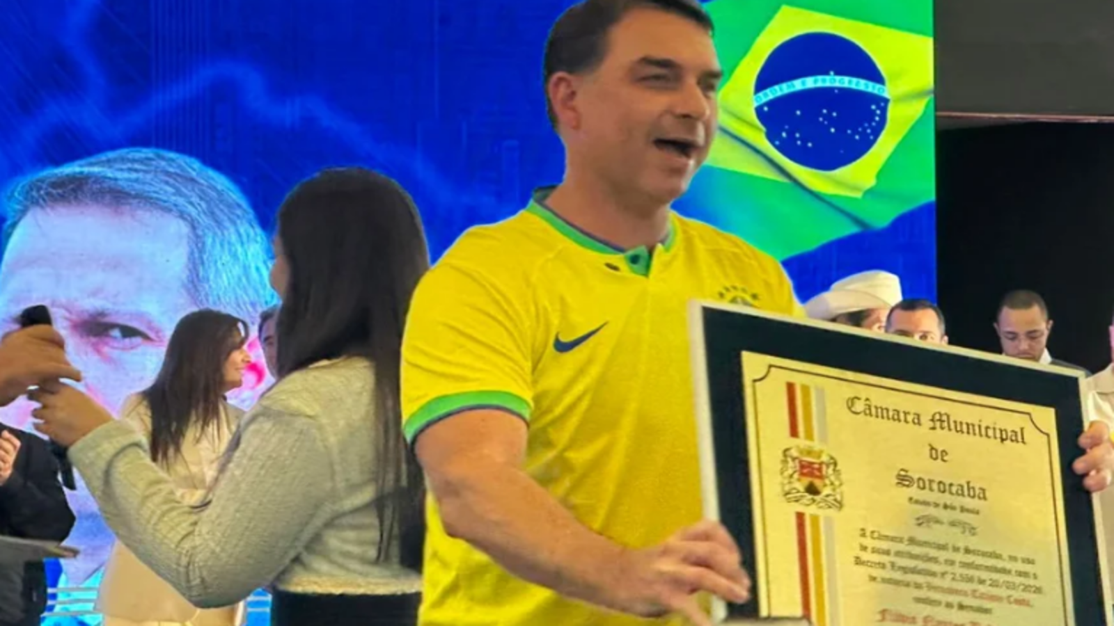 Man in a yellow Brazil jersey holds a large framed certificate on stage with a Brazilian flag in the background