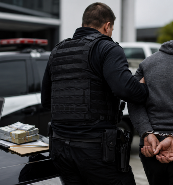 Police officer in tactical vest escorts a handcuffed suspect toward a police car on the hood are stacks of cash several laptops and multiple smartphones
