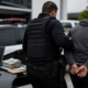 Police officer in tactical vest escorts a handcuffed suspect toward a police car on the hood are stacks of cash several laptops and multiple smartphones