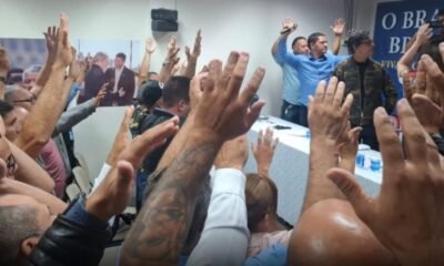 Crowd with raised hands facing a panel at a meeting blue banner visible in the background