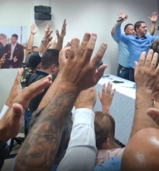 Crowd with raised hands facing a panel at a meeting blue banner visible in the background