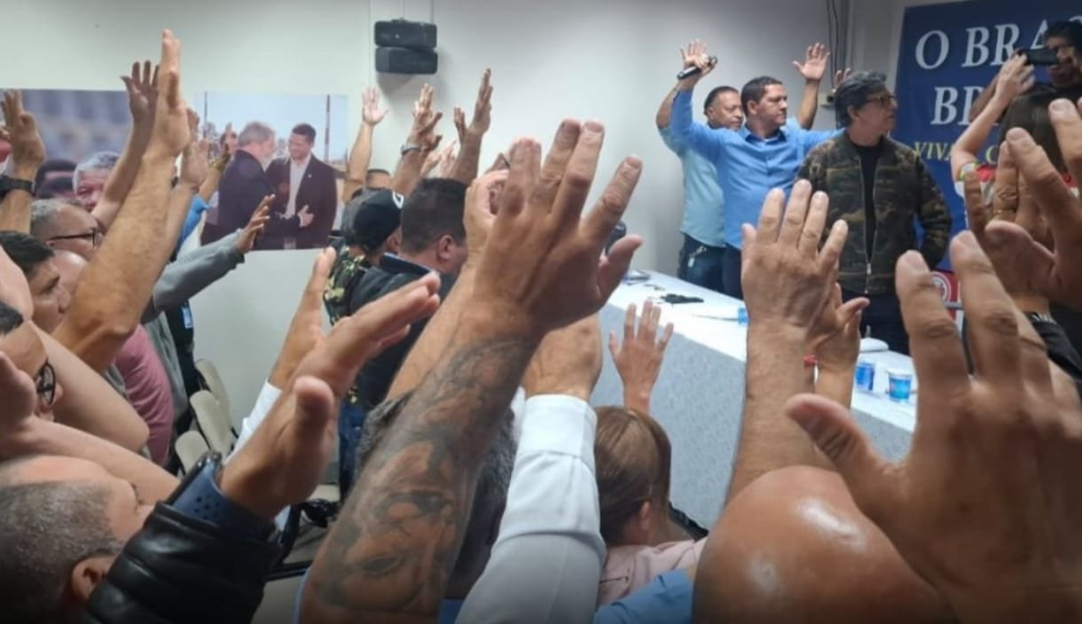 Crowd with raised hands facing a panel at a meeting blue banner visible in the background